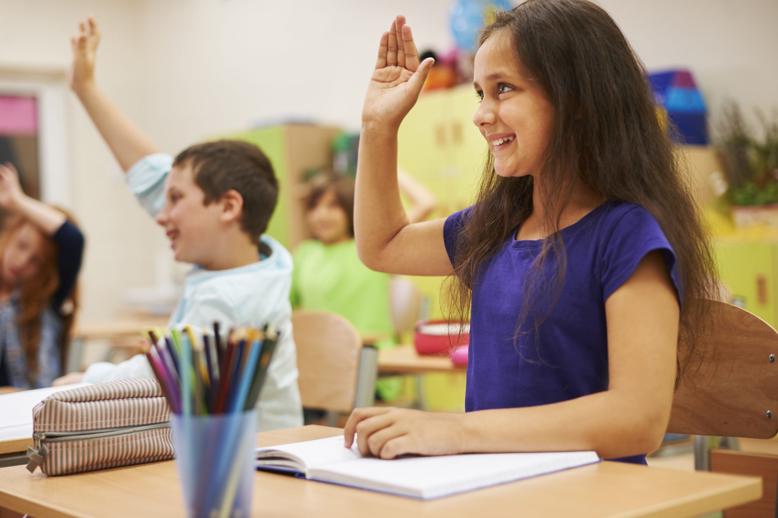 Menina sentada sorri com a mão levantada participando da aula