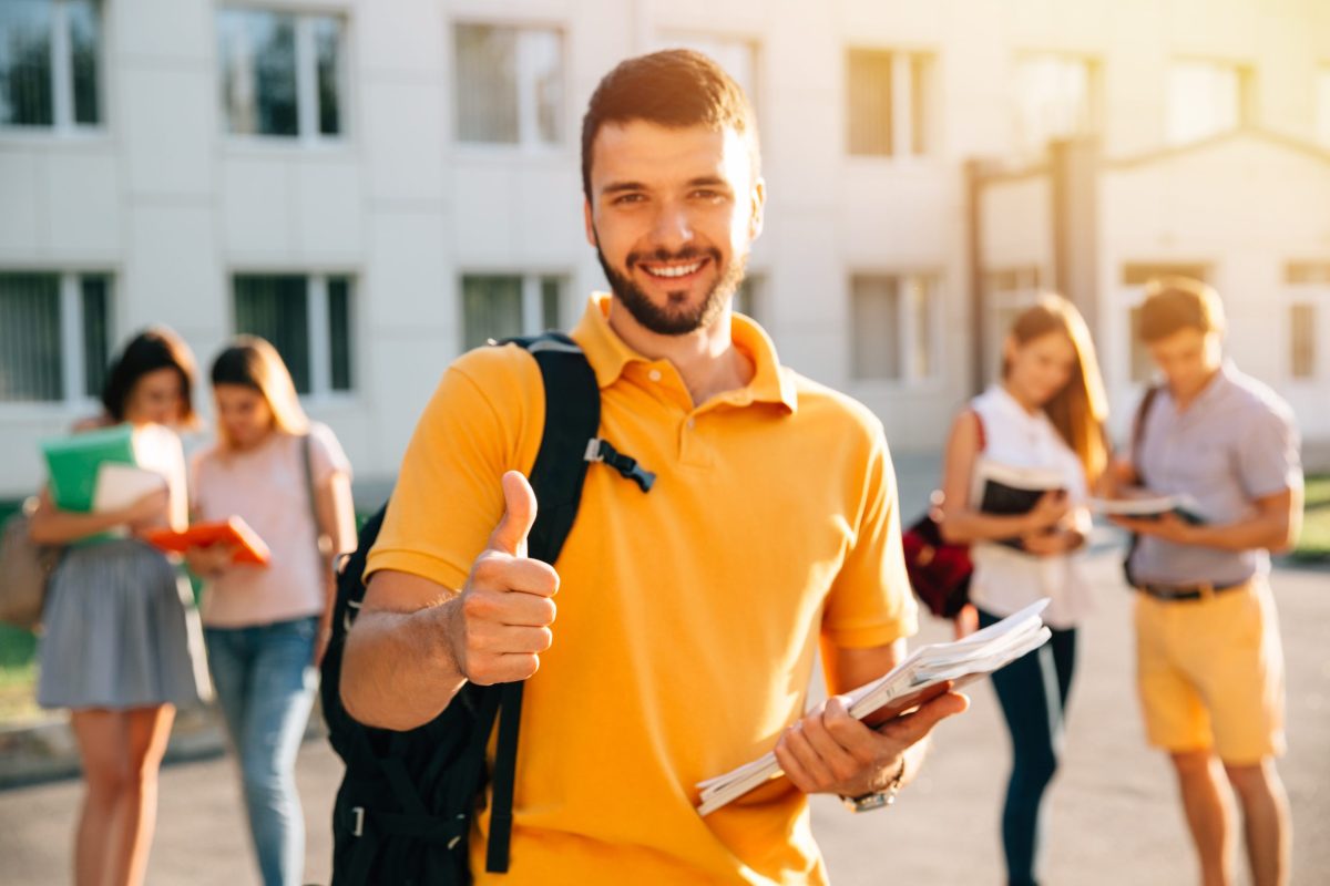 Jovem universitário sorri e faz sinal de positivo parado no campus carregando mochila e cadernos nas mãos