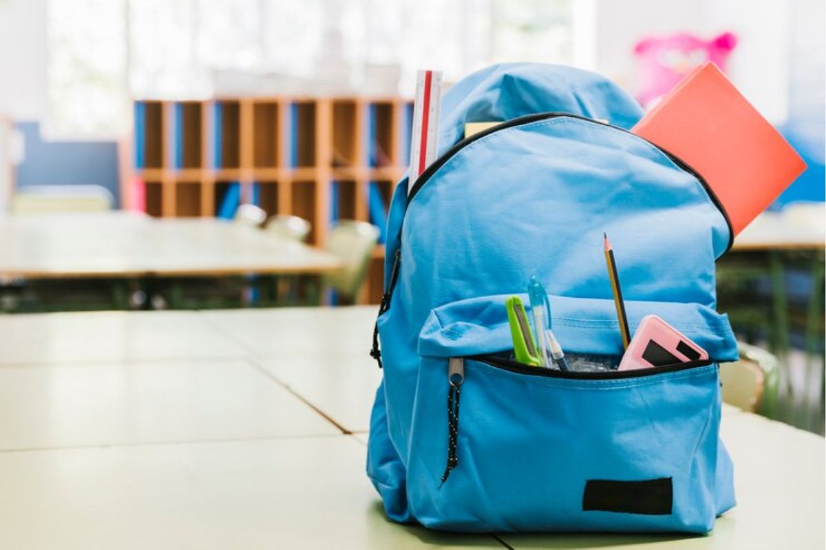 Uma mochila azul está em cima de uma mesa na escola, é possível ver alguns materiais nela