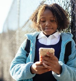 Uma menina está de casaco, encostada na grade e segura um celular. Ela está conferindo o Boletim Online 2024 PR