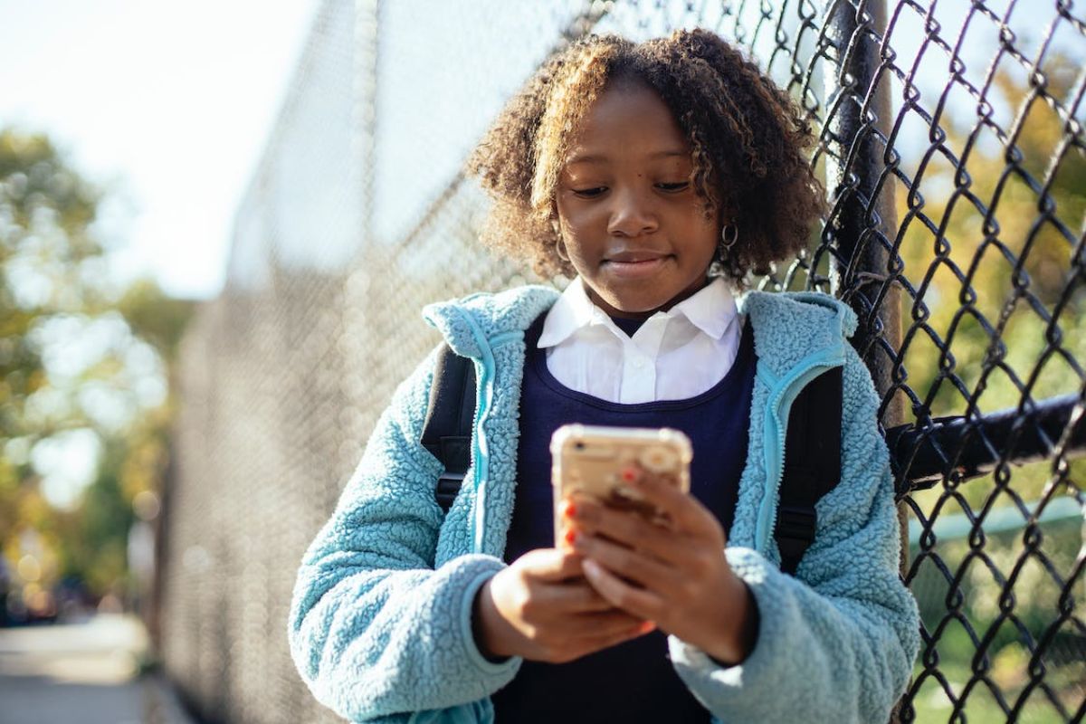 Uma menina está de casaco, encostada na grade e segura um celular. Ela está conferindo o Boletim Online 2024 PR