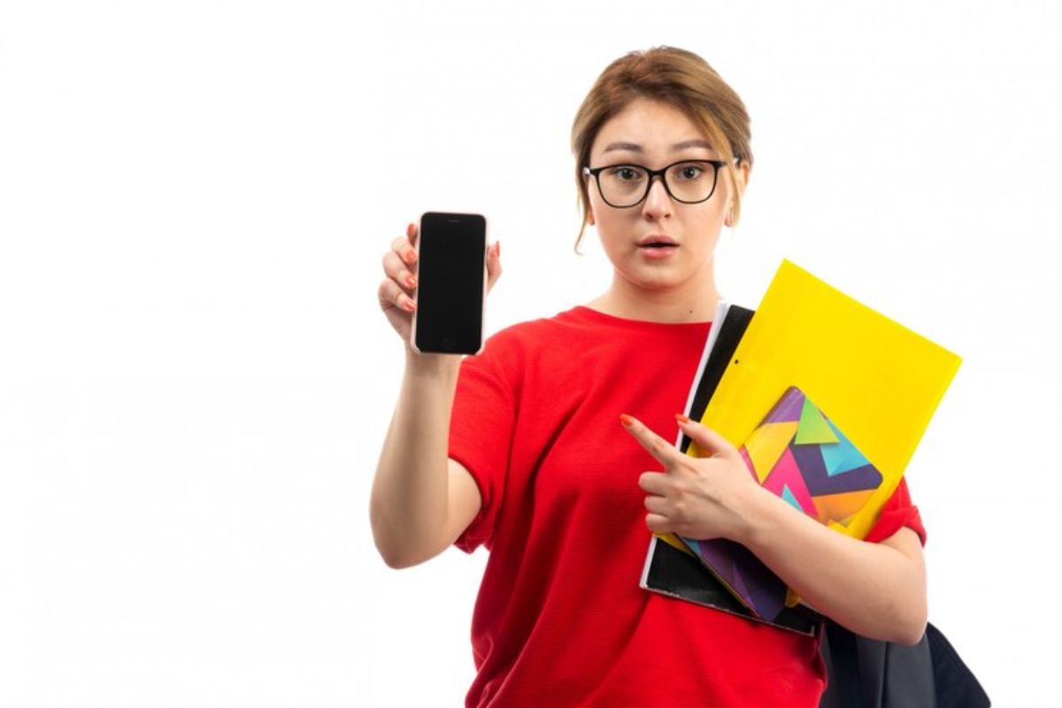 Uma mulher segura seu material escolar em um braço e aponta para o celular que está em outra mão