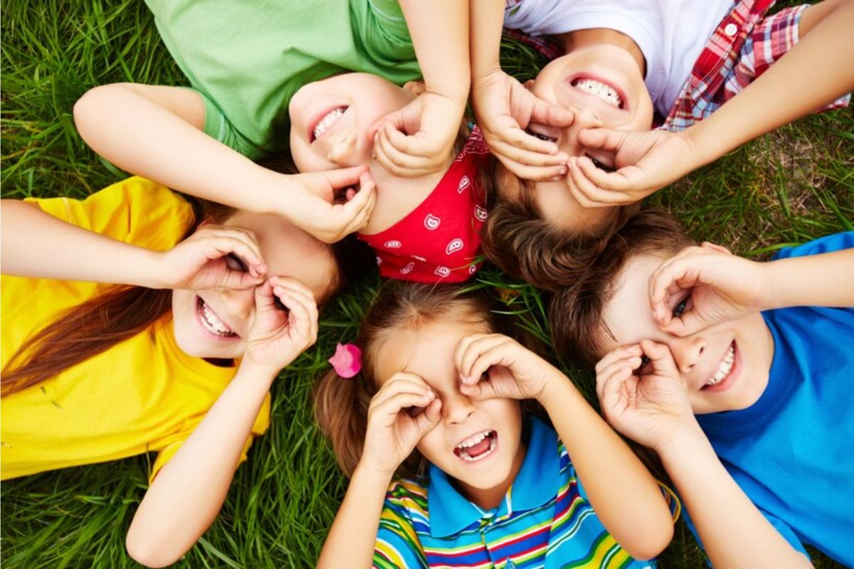 Cinco crianças deitas na grama fazem sinal de binóculos com as mãos nos rostos